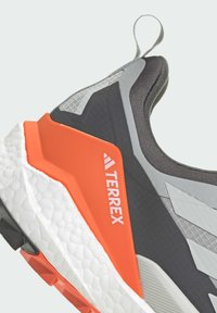 Szare i czarne buty sportowe z pomarańczowymi akcentami, teksturowana siateczkowa cholewka oraz gruba biała podeszwa. Zawierają logo oraz zorganizowane elementy designu.