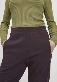 Persoon in een ribbelige olijfgroen lange mouw top die in hoge paarse broek is gestopt, met handen in de zakken, tegen een lichte achtergrond.