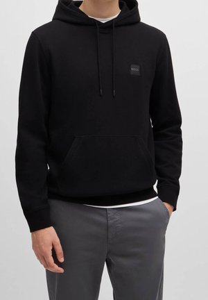 Homme portant un sweat à capuche noir avec le logo BOSS sur la poitrine et un pantalon gris, mains détendues le long du corps sur un fond uni.