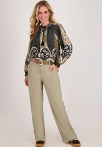 Vrouw die glimlacht in een gestreepte blouse met lange mouwen, beige wijde broek met riem en donkere bruine schoenen, staande tegen een neutrale achtergrond.