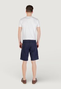 Vista posteriore di un uomo che indossa una t-shirt bianca in cotone, pantaloni corti in cotone blu navy e scarpe in pelle marrone. Design semplice senza motivi visibili.