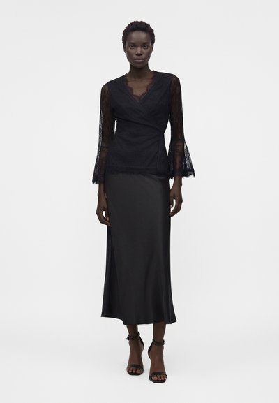Μαύρο δαντελένιο πουκάμισο με μακριά μανίκια, συνδυασμένο με μαύρη σατέν midi φούστα και μαύρα πέδιλα με λουράκι στον αστράγαλο σε ένα μοντέλο που στέκεται.