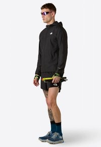 Svart lättviktsjacka med huva, svarta shorts, blå löparskor och lila solglasögon. Håller i vandringsstavar med neonfärgade detaljer.