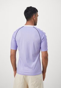 Uomo di spalle che indossa una maglietta a maniche corte con strisce verticali viola e bianche e pantaloni beige chiaro, su sfondo uniforme.