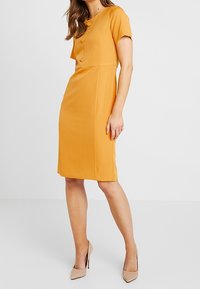 Vestido amarillo mostaza con mangas cortas y escote redondo, que presenta tres botones en la parte delantera. Diseño ajustado y de longitud hasta la rodilla.
