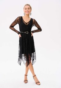 Zwarte kanten jurk met lange semi-doorzichtige mouwen en een V-halslijn. Hoge-lage zoomlijn. Gestyled met een zwarte riem en bruine sandalen met bandjes.