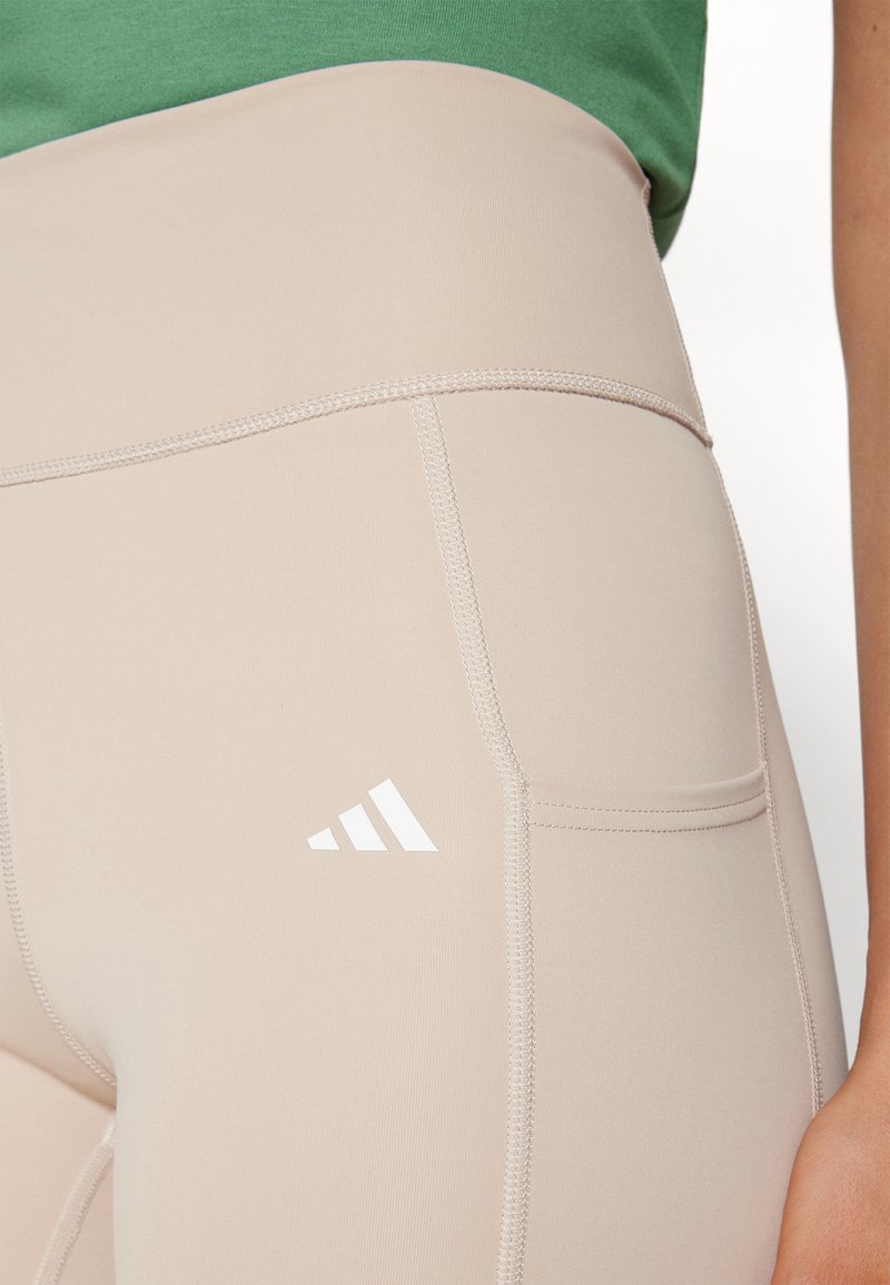 Leggings de talle alto en color beige, fabricados en un material elástico, con un logo blanco y detalles de costura, y bolsillos laterales para mayor funcionalidad.