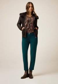 Chaqueta bomber negra con ribete de piel, blusa de capas con volantes en estampado floral oscuro, pantalones rectos en color teal, y botas de tobillo marrones.