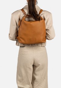 Un sac à dos en cuir marron avec une surface texturée, une forme arrondie, des bretelles réglables et une étiquette rectangulaire, porté sur le dos d'une personne.