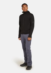 Zwarte zip-up hoodie met hoge kraag, gecombineerd met lichtgrijze broeken en zwart schoeisel. Stof lijkt glad; minimale branding zichtbaar op de mouw.