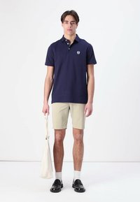 Jūros mėlynumo polo marškiniai su trumpomis rankovėmis ir apykakle, smėlio spalvos šortai, juodi slip-on batai ir kreminės spalvos rankinė. Minimalistinis dizainas, gerai prigludęs.