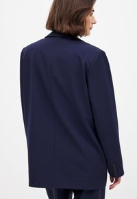 Navy blazer with a tailored fit, featuring a notched lapel, long sleeves, and button accents on the cuffs. Smooth fabric texture and sleek design.