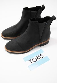 Bottines Chelsea en daim noir avec des panneaux latéraux élastiques et une semelle en caoutchouc plate. Présentent un liseré marron cousu et une étiquette avec le logo TOMS sur fond bleu.