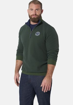 Bearded man wearing a dark green quarter-zip pullover with logo on chest and dark blue jeans, standing against a plain background.