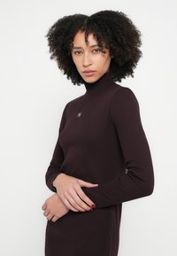 Burgunderfarget fotsid genserkjole med ribbestruktur, lange ermer og liten logo på brystet. Den tettsittende silhuetten fremhever formen.