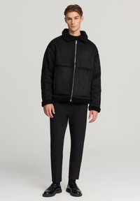 Giacca nera in shearling sintetico con chiusura a zip, ampio colletto e tasche laterali. Abbinata a pantaloni neri aderenti e scarpe di pelle.