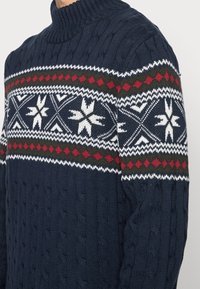 Granatowy sweter w warkocz z białym i czerwonym wzorem diamentów oraz płatków śniegu na klatce piersiowej i górnych ramionach, noszony przez osobę.