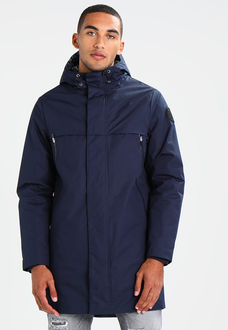 Cappotto impermeabile blu navy con cappuccio, dotato di tasche anteriori con zip, un design a cucitura doppia e un badge testurizzato sulla manica.