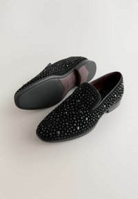 Schwarze Slipper mit texturierter Oberfläche, verziert mit pyramidenförmigen Nieten; verfügt über eine glatte schwarze Sohle und ein dunkelburgunderfarbenes Innenleben.