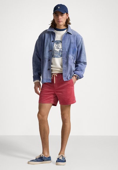 Plava traper jakna preko bijelog duksera s grafikom, crvene kordne kratke hlače i plave tenisice s svijetlim vezicama. Opušteni ljetni outfit.