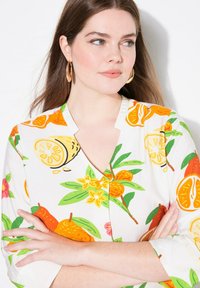 Hvid bluse med livligt frugtmønster, der indeholder appelsiner, citroner og blomster. Bemærkelsesværdige detaljer inkluderer en V-udskæring og trekvartærmer.