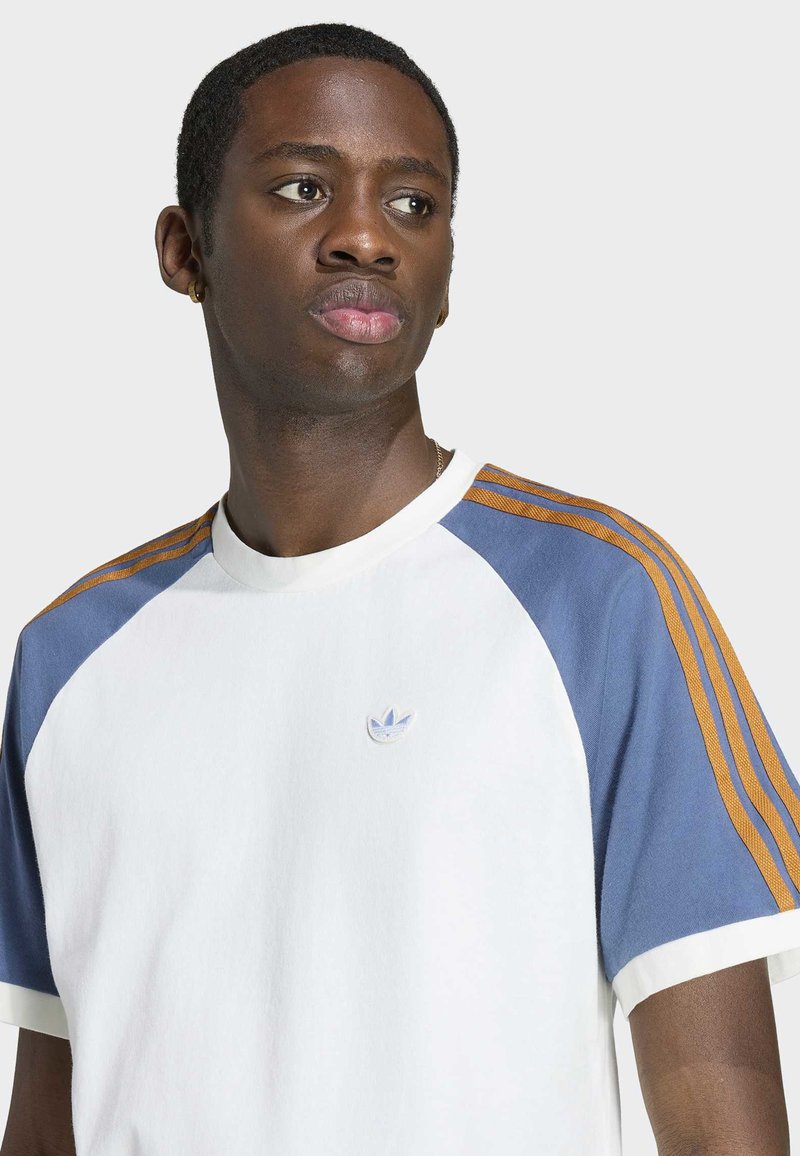 Junger Mann schaut seitlich, trägt ein weißes T-Shirt mit blauen Ärmeln und braunen Streifen sowie das Adidas-Logo auf der Brust vor neutralem Hintergrund.