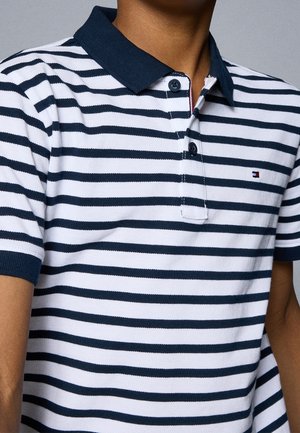 Persona con una camiseta polo con rayas horizontales en azul marino y blanco, con cuello azul marino y pequeño logo en el pecho izquierdo.