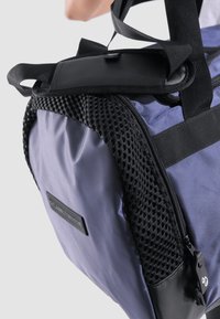 Bolso púrpura con detalles de malla, correas negras y una superficie texturizada. Presenta un cierre de cremallera y un parche con el logo en el costado.