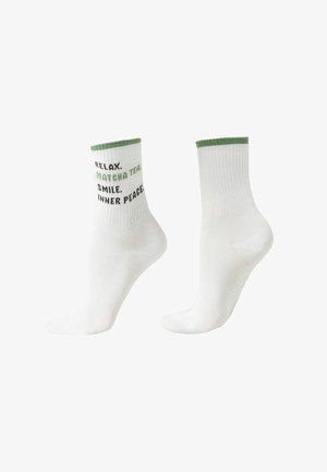 Witte enkelsokken met groene rand, waarvan één tekst toont: "ONTSPAN. MATCHA THEE. GLIMLACH. INNERLIJKE RUST" in zwarte en groene letters.