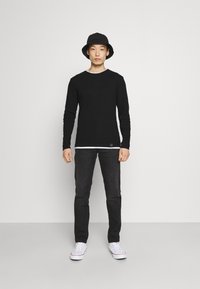 Chemise noire à manches longues par-dessus une chemise blanche, jeans noirs et baskets blanches avec un chapeau seau noir. Conception simple et ajustée.