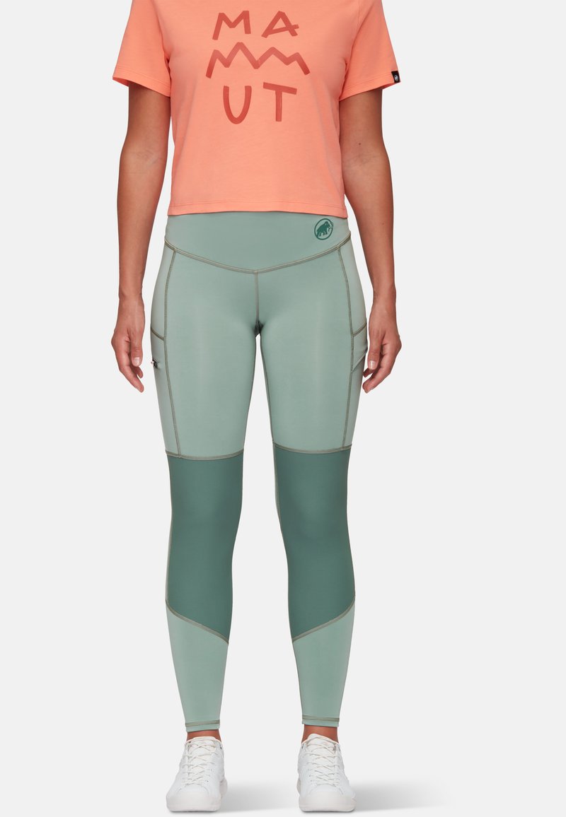 Leggings verde chiaro con pannelli verde scuro, caratterizzati da cuciture a contrasto e un logo in vita, abbinati a una maglietta arancione corta.
