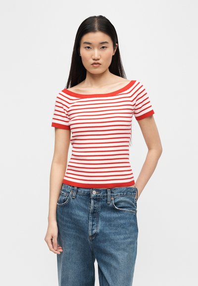 Νεαρή γυναίκα με μακριά σκούρα μαλλιά, που φοράει κόκκινη και λευκή ριγέ κοντομάνικη μπλούζα και ψηλόμεσα μπλε τζιν, στέκεται μπροστά σε μια απλή φόντο.