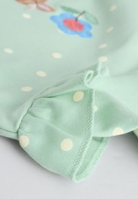 Világoszöld szövet fehér pöttyökkel, fodros széllel, és egy kék virággal, amelynek piros középpontja és zöld levelei vannak rányomtatva.