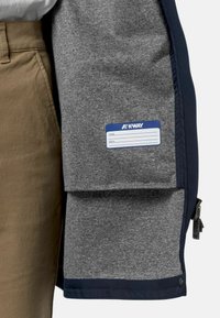 Grijze jas met marineblauwe accenten, voorzien van een gladde textuur, een rits aan de zijkant en een binnentag voor naam en e-mailadres.