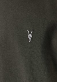 Tumma vihreä kangas, jossa on pehmeä rakenne, sisältää pienen valkoisen brodeeratun vuohenkallon logon vasemmalla ylhäällä.