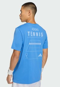 Niebieski bawełniany t-shirt z napisem "ADIDAS TENNIS" i grafiką kortu tenisowego na plecach. Krótkie rękawy, luźny krój i lekka tekstura.