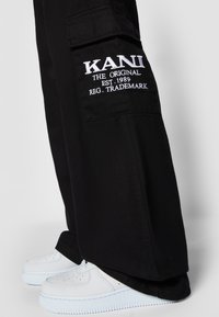 Black cargo pants with a large side pocket featuring white embroidered text. Paired with white sneakers showing textured surfaces.
