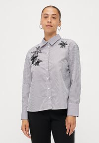 Striped button-up shirt in white and navy. Features black embroidered floral accents on the chest, with a collar and long sleeves.