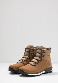 Bottes de randonnée marron avec une tige en tissu texturé, des semelles en caoutchouc noir et des lacets gris. Elles présentent un col rembourré et un logo sur le côté.