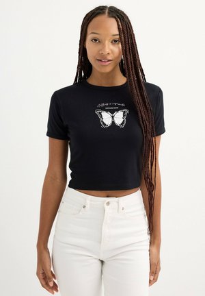 Camiseta corta negra con un gráfico de mariposa blanca y texto, hecha de una tela suave. Combinada con jeans blancos de tiro alto.