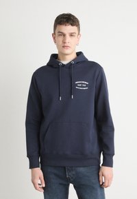Marineblå hettegenser med snor, frontlomme og hvit trykt tekst. Stoffet har en glatt tekstur, og ermene er ribbestrikkede.