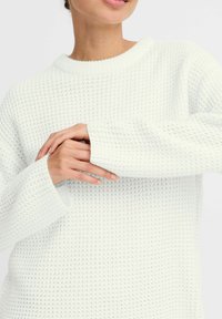 Witte gebreide trui met een gestructureerd patroon, brede mouwen en een ronde halslijn. De stof is zacht en licht chunky.