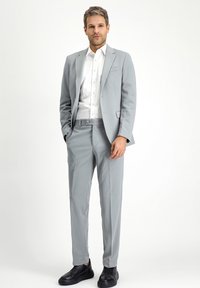 Heller grauer Anzug aus glattem Stoff, kombiniert mit einem weißen Hemd. Schwarze Schuhe runden den Look ab, mit moderner Passform und schickem Design.
