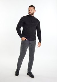 Maglione nero a maglia con collo alto e zip argentata, abbinato a jeans scuri e scarpe nere, in piedi davanti a uno sfondo bianco.
