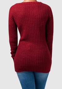 Maglione rosso lavorato a maglia con un design testurizzato e a motivi. Maniche lunghe e scollo rotondo. Orlo e polsini a costine. Indossato con jeans blu.
