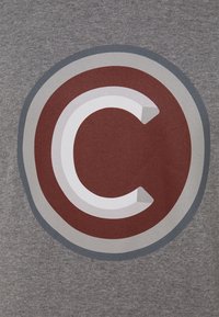 Vit bokstav C inuti en röd cirkel med ljusgrå och mörkgrå ringar på en texturerad grå tygbakgrund.