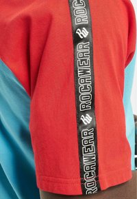 Raudonas ir mėlynas trumpomis rankovėmis marškinėliai su juodu logotipo juostele, ant kurios yra užrašas "ROC WEAR", einantis per pečių siūlę. Pagaminta iš medvilnės mišinio.