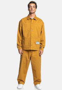 Katoenen werkkledingjas en -broek in mosterdgeel. Voorzien van een knoopsluiting aan de voorkant, twee voorkruiszakken en een logo-opmerking op de jas.