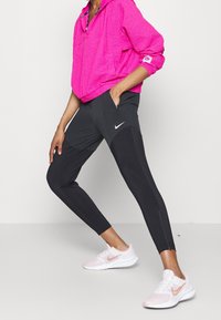 Osoba w jasnoróżowej kurtce i czarnych legginsach Nike stoi z jedną ręką w kieszeni, nosząc różowo-białe sneakersy Nike.