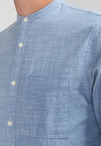 Helles blaues Hemd mit Kragen aus strukturiertem Stoff, ausgestattet mit einer Brusttasche und weißen Knöpfen entlang der Vorderseite mit einem dezenten Muster.